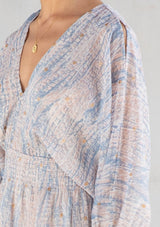 [Color: Sky Natural] A detail image of a brunette model wearing a boho mini dress with an abstract print, split sleeves, a v neckline, smocked waist, and a tassel tie back detail. 