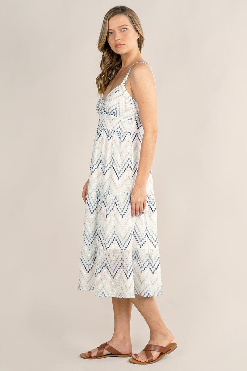 [Color: White/Blue] A side facing image of a blonde model wearing a white and blue chevron pattern midi dress with all over eyelet details. Featuring a sleeveless design, a v-neckline, smocked back, button front top, and flowy tiered skirt. 