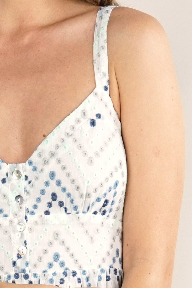 [Color: White/Blue] A detail image of a blonde model wearing a white and blue chevron pattern midi dress with all over eyelet details. Featuring a sleeveless design, a v-neckline, smocked back, button front top, and flowy tiered skirt. 