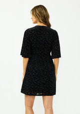 [Color: Black] A back facing image of a brunette model wearing a black velvet burnout mini dress with half sleeves, a v neckline, and an elastic empire waist. 