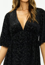 [Color: Black] A detail image of a brunette model wearing a black velvet burnout mini dress with half sleeves, a v neckline, and an elastic empire waist. 