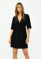 [Color: Black] A front facing image of a brunette model wearing a black velvet burnout mini dress with half sleeves, a v neckline, and an elastic empire waist. 