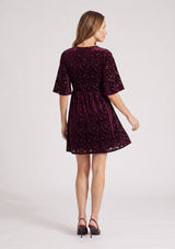 [Color: Merlot] A back facing image of a blonde model wearing a burgundy velvet burnout mini dress with half sleeves, a v neckline, and an elastic empire waist. 