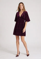 [Color: Merlot] A front facing image of a blonde model wearing a burgundy velvet burnout mini dress with half sleeves, a v neckline, and an elastic empire waist. 