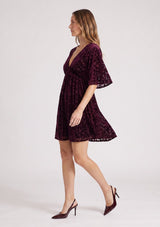 [Color: Merlot] A side facing image of a blonde model wearing a burgundy velvet burnout mini dress with half sleeves, a v neckline, and an elastic empire waist. 