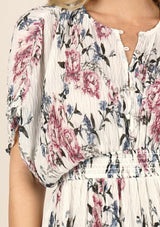[Color: Ivory Orchid] A detail image of a blonde model wearing a pink and blue floral print mini dress with a button front top, short dolman sleeves, and a smocked waistline. 