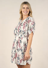 [Color: Ivory Orchid] A side facing image of a blonde model wearing a pink and blue floral print mini dress with a button front top, short dolman sleeves, and a smocked waistline. 