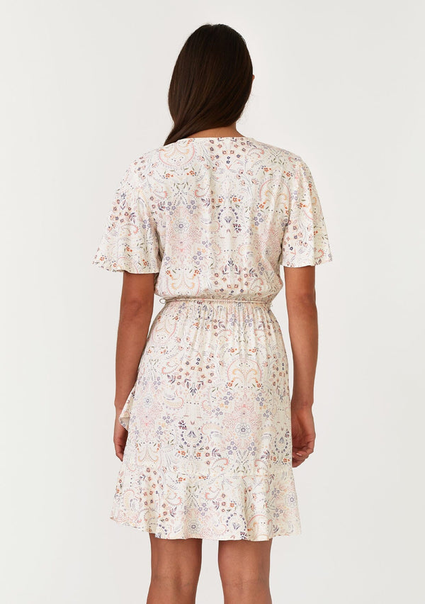 [Color: Natural/Peach] A back facing image of a brunette model wearing a bohemian ivory and pink floral print mini dress. With short flutter sleeves, a surplice v neckline, an elastic waist, a tassel tie waist belt, and a ruffled hemline. 