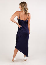 [Color: Navy] A back facing image of a blonde woman wearing a sexy and chic silky asymmetrical maxi dress in blue with subtle dot details, a v neckline, and spaghetti straps. 