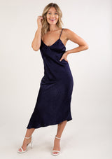 [Color: Navy] A front facing image of a blonde woman wearing a sexy and chic silky asymmetrical maxi dress in blue with subtle dot details, a v neckline, and spaghetti straps. 