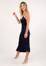 [Color: Navy] A side facing image of a blonde woman wearing a sexy and chic silky asymmetrical maxi dress in blue with subtle dot details, a v neckline, and spaghetti straps. 