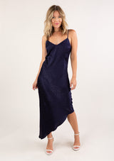 [Color: Navy] A blonde woman wearing a sexy and chic silky asymmetrical maxi dress in blue with subtle dot details, a v neckline, and spaghetti straps. 