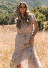 [Color: Ivory/Taupe] A close up front facing image of a blonde model standing in a field wearing an ivory and brown animal print boho maxi dress. With short flutter sleeves, a v neckline, a smocked elastic waist with tassel tie accent, and a flowy high low skirt and tiered skirt.