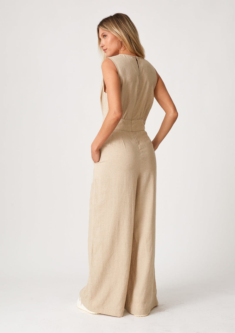 [Color: Wheat] A back facing image of a blonde model wearing linen blend full length jumpsuit. With a wide leg, side pockets, a v neckline, and a back zip closure.