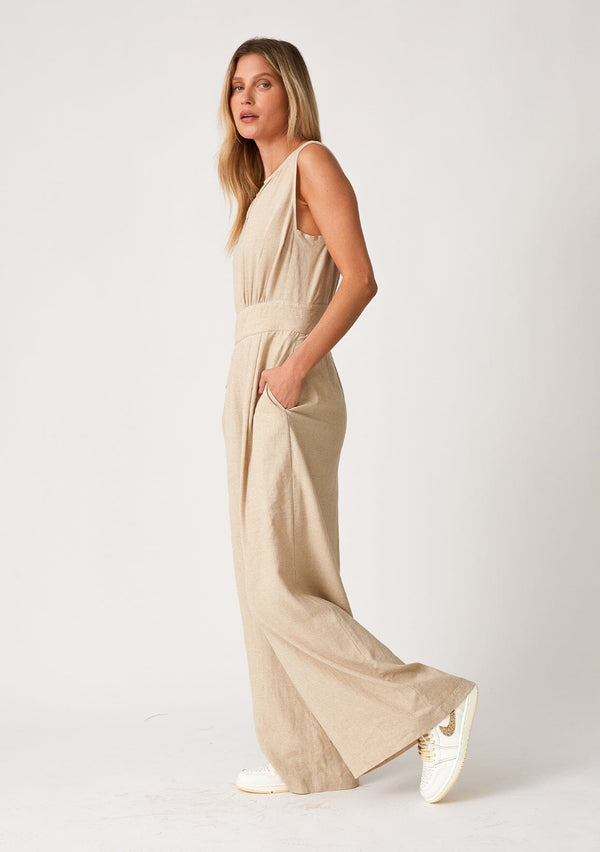 [Color: Wheat] A side facing image of a blonde model wearing linen blend full length jumpsuit. With a wide leg, side pockets, a v neckline, and a back zip closure.