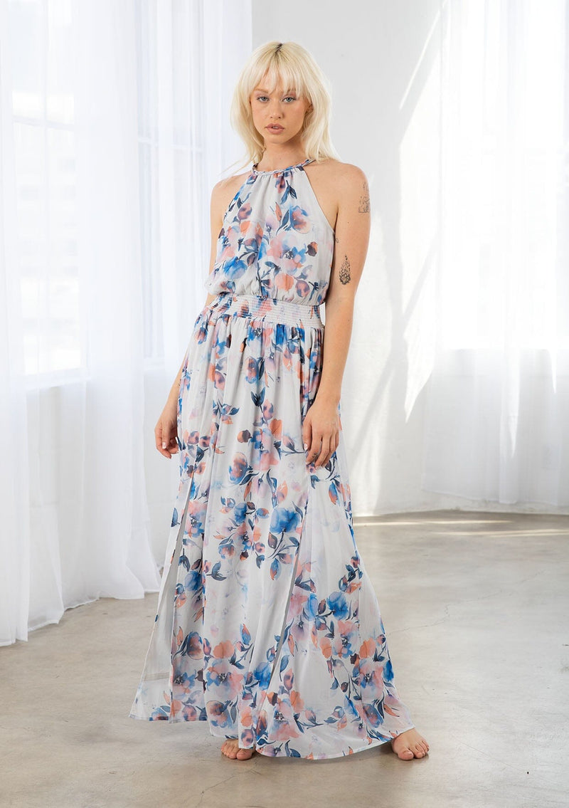 [Color: Ivory/Blue] A model wearing a dreamy sheer floral maxi dress with a braided trimmed halter neckline, a smocked elastic waist, a sexy side slit, and a back keyhole with braided rope tie. 