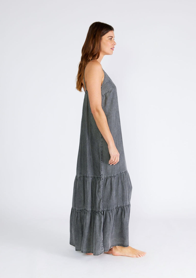 [Color: Ash Grey Wash] A side facing image of a brunette model wearing an ash grey wash sleeveless maxi dress made from Tencel. With adjustable spaghetti straps, a v neckline, and a long flowy tiered silhouette. 