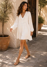 [Color: Vanilla/Sand] A woman wearing a white long-sleeve embroidered dress walks outdoors on a sunlit stone path beside a beige stucco wall and potted plants.