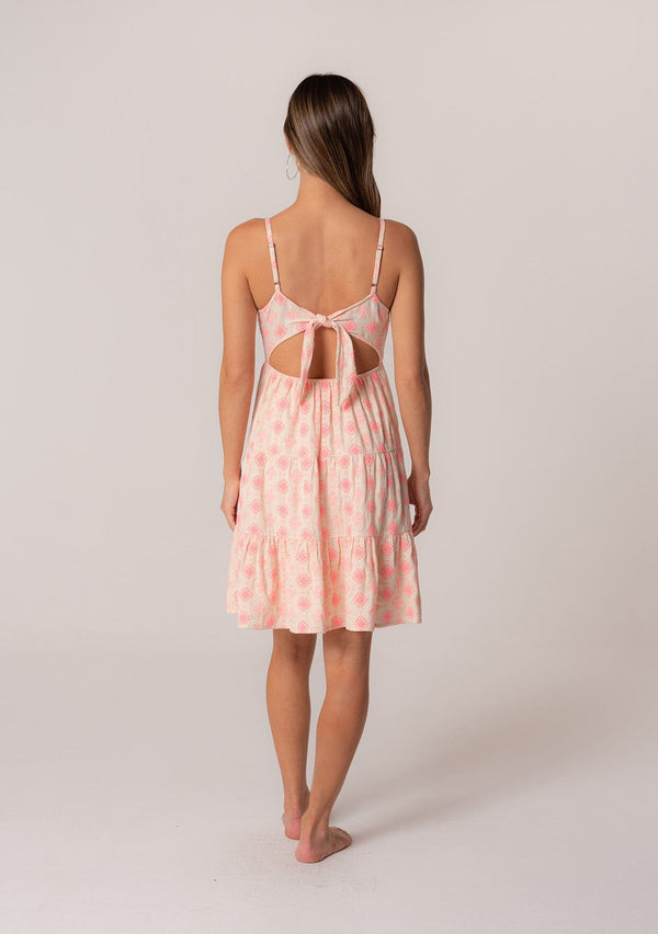 [Color: Natural/Pink] A back facing image of a brunette model wearing a sleeveless cotton mini dress in pink embroidery. With adjustable spaghetti straps, a square neckline, a tiered silhouette, an empire waist, an open back detail, and an adjustable tie back. 