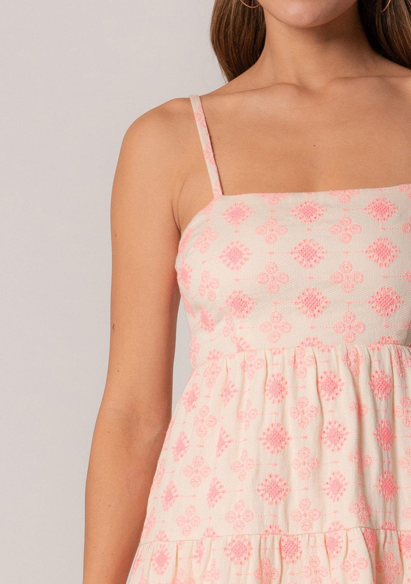 [Color: Natural/Pink] A close up front facing image of a brunette model wearing a sleeveless cotton mini dress in pink embroidery. With adjustable spaghetti straps, a square neckline, a tiered silhouette, an empire waist, an open back detail, and an adjustable tie back. 