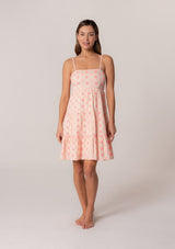[Color: Natural/Pink] A front facing image of a brunette model wearing a sleeveless cotton mini dress in pink embroidery. With adjustable spaghetti straps, a square neckline, a tiered silhouette, an empire waist, an open back detail, and an adjustable tie back. 