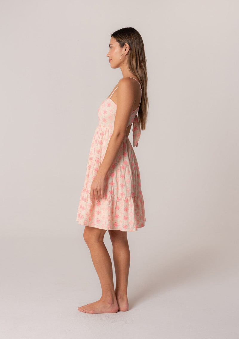 [Color: Natural/Pink] A side facing image of a brunette model wearing a sleeveless cotton mini dress in pink embroidery. With adjustable spaghetti straps, a square neckline, a tiered silhouette, an empire waist, an open back detail, and an adjustable tie back. 