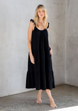 [Color: Black] A model wearing a classic black maxi dress designed in soft cotton gauze. With a scooped neckline, adjustable ruffled straps, and side pockets.