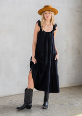 [Color: Black] A model wearing a classic black maxi dress designed in soft cotton gauze. With a scooped neckline, adjustable ruffled straps, and side pockets.