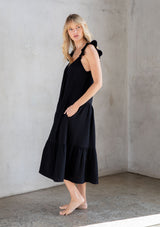 [Color: Black] A model wearing a classic black maxi dress designed in soft cotton gauze. With a scooped neckline, adjustable ruffled straps, and side pockets.