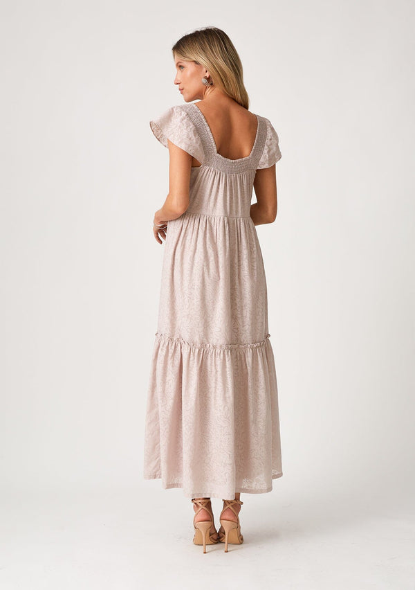 [Color: Dusty Lavender] A back facing image of a blonde model wearing a dreamy bohemian cotton blend maxi dress in a light lavender. With a smocked square neckline, short flutter cap sleeves, and a ruffle trimmed tiered hemline.