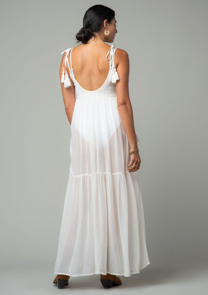 [Color: Ivory] A back facing image of a brunette model wearing an ivory sheer maxi cover up beach dress. With adjustable tassel tie spaghetti tank top straps, a deep v neckline, a side lace up detail, and a lace trim top.