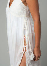 [Color: Ivory] A close up side facing image of a brunette model wearing an ivory sheer maxi cover up beach dress. With adjustable tassel tie spaghetti tank top straps, a deep v neckline, a side lace up detail, and a lace trim top.