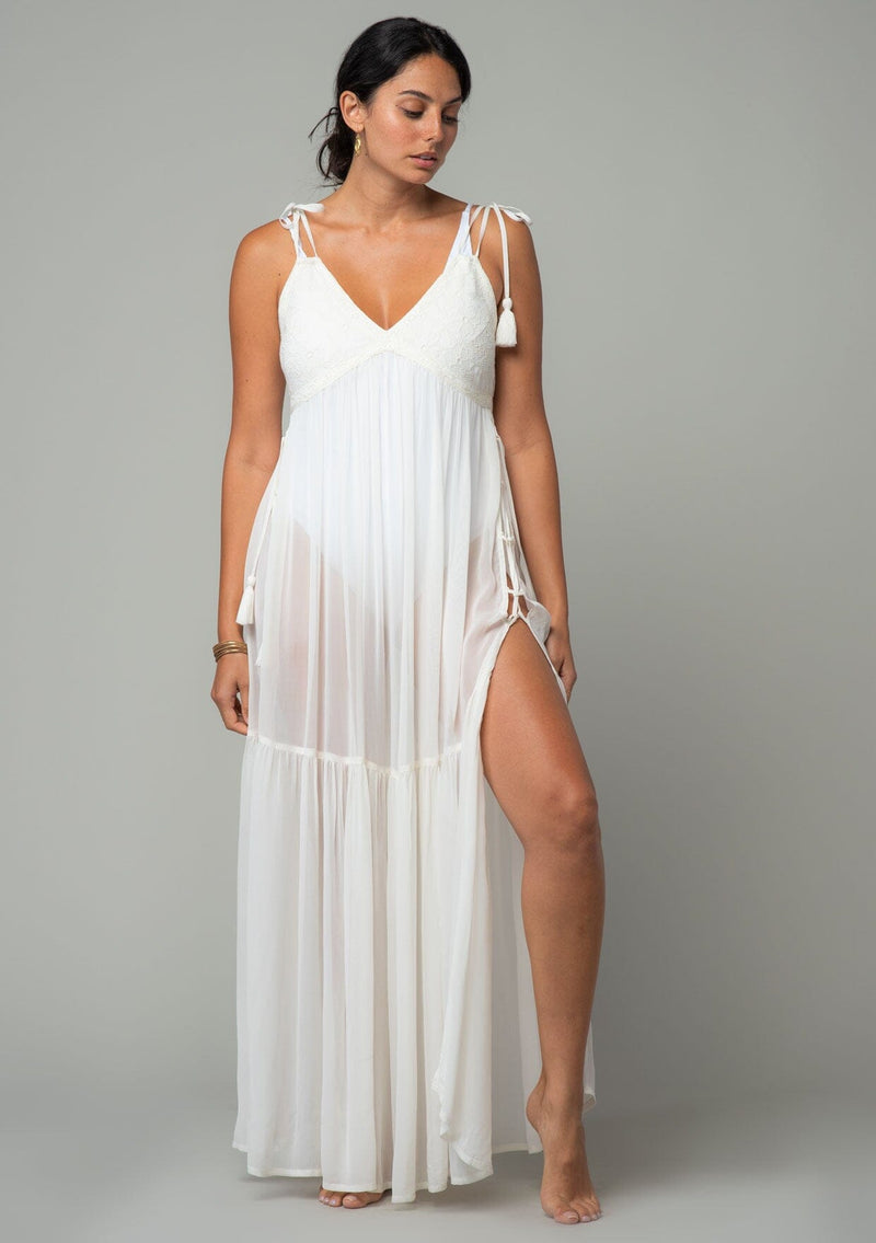 [Color: Ivory] A front facing image of a brunette model wearing an ivory sheer maxi cover up beach dress. With adjustable tassel tie spaghetti tank top straps, a deep v neckline, a side lace up detail, and a lace trim top.
