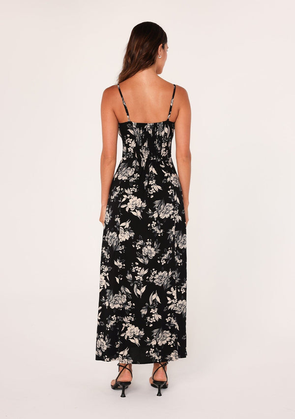[Color: Black/Natural] A back facing image of a brunette model wearing a special occasion boho maxi dress in a black and off white floral print. With adjustable spaghetti straps, a drawstring neckline with tassel ties, a half smocked elastic bodice at the back, and a long flowy skirt with a slit. 