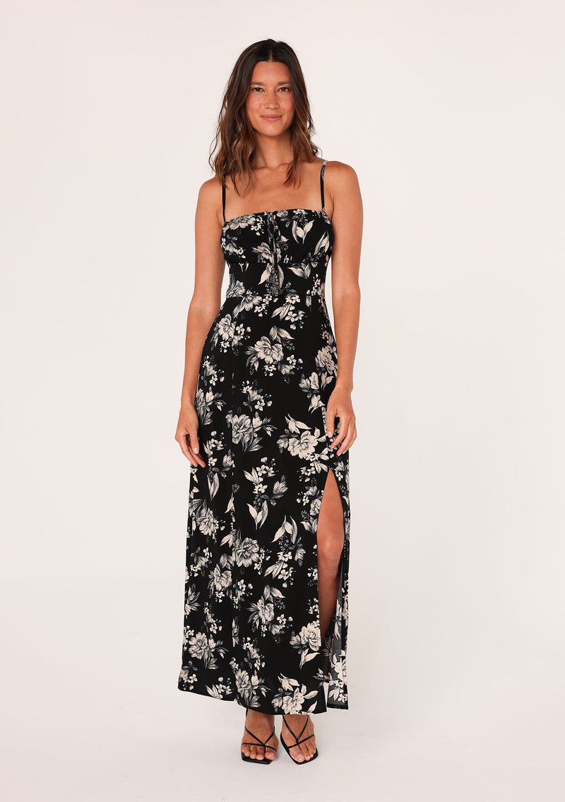 [Color: Black/Natural] A front facing image of a brunette model wearing a special occasion boho maxi dress in a black and off white floral print. With adjustable spaghetti straps, a drawstring neckline with tassel ties, a half smocked elastic bodice at the back, and a long flowy skirt with a slit. 