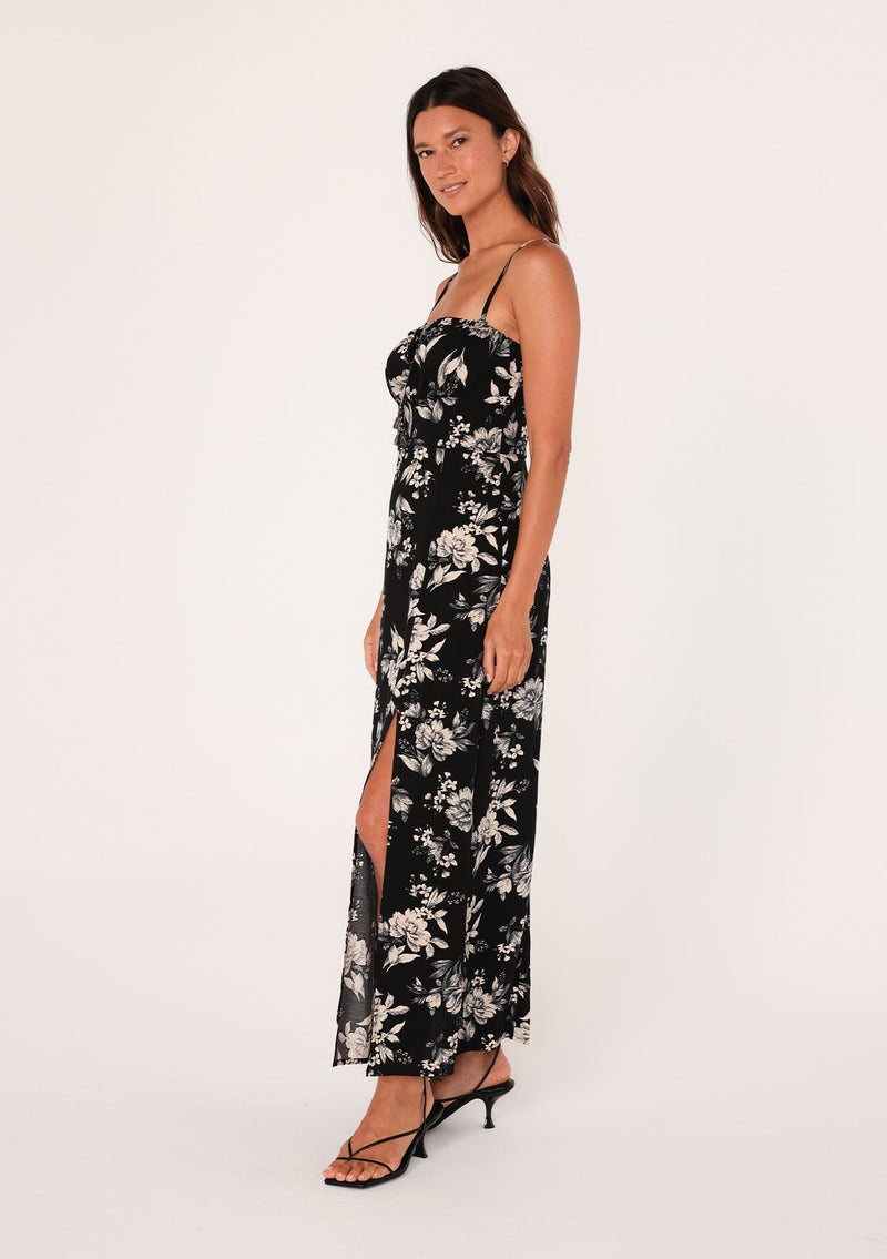 [Color: Black/Natural] A side facing image of a brunette model wearing a special occasion boho maxi dress in a black and off white floral print. With adjustable spaghetti straps, a drawstring neckline with tassel ties, a half smocked elastic bodice at the back, and a long flowy skirt with a slit. 