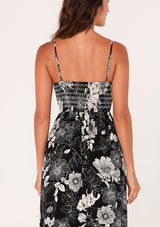[Color: Black/White] A close up back facing image of a brunette model wearing a sleeveless maxi dress in a black and white floral print. With adjustable spaghetti straps, a square neckline, a slim fit smocked bodice, a decorative button front top, side pockets, and a long flowy skirt. 