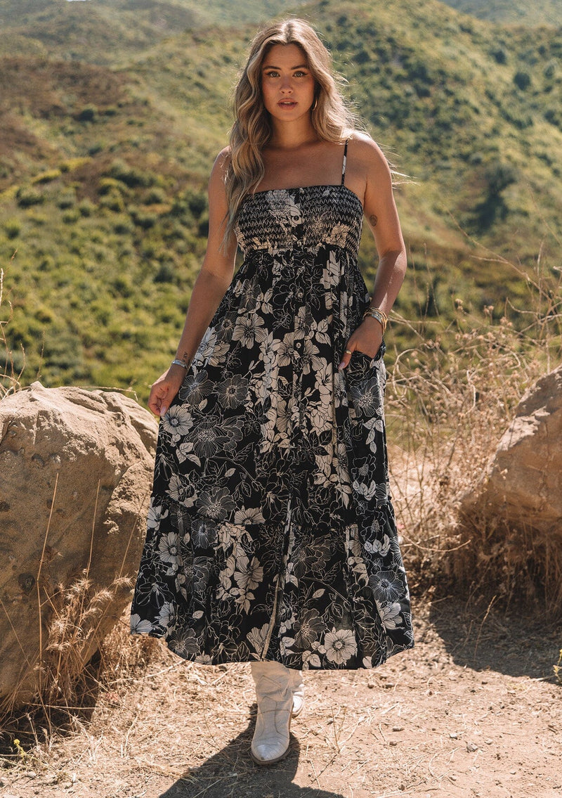 [Color: Black/White] A front facing image of a blonde model standing outside wearing a sleeveless maxi dress in a black and white floral print. With adjustable spaghetti straps, a square neckline, a slim fit smocked bodice, a decorative button front top, side pockets, and a long flowy skirt.