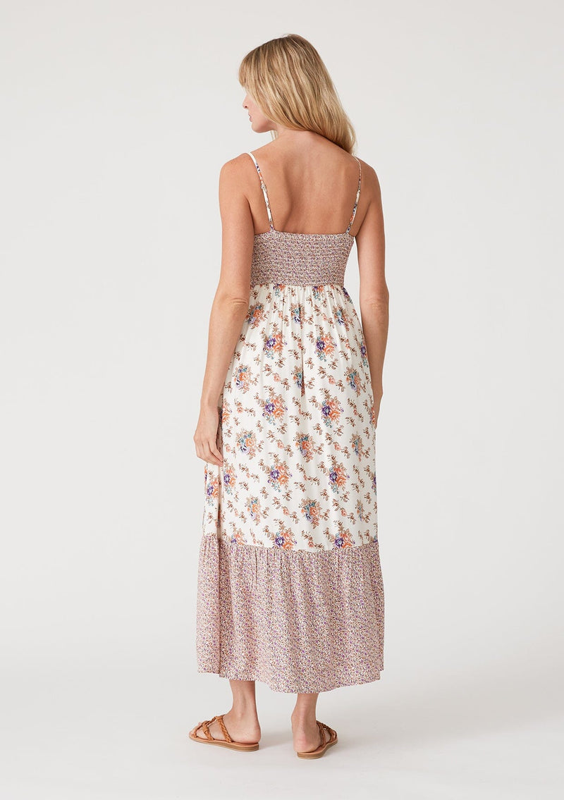 [Color: Natural/Purple] A back facing image of a blonde model wearing a best selling bohemian maxi dress in an off white and purple floral border print. With adjustable spaghetti straps, a square neckline, a smocked fitted bodice, a flowy tiered skirt, side pockets, and a button front. 