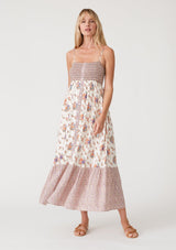 [Color: Natural/Purple] A front facing image of a blonde model wearing a best selling bohemian maxi dress in an off white and purple floral border print. With adjustable spaghetti straps, a square neckline, a smocked fitted bodice, a flowy tiered skirt, side pockets, and a button front. 