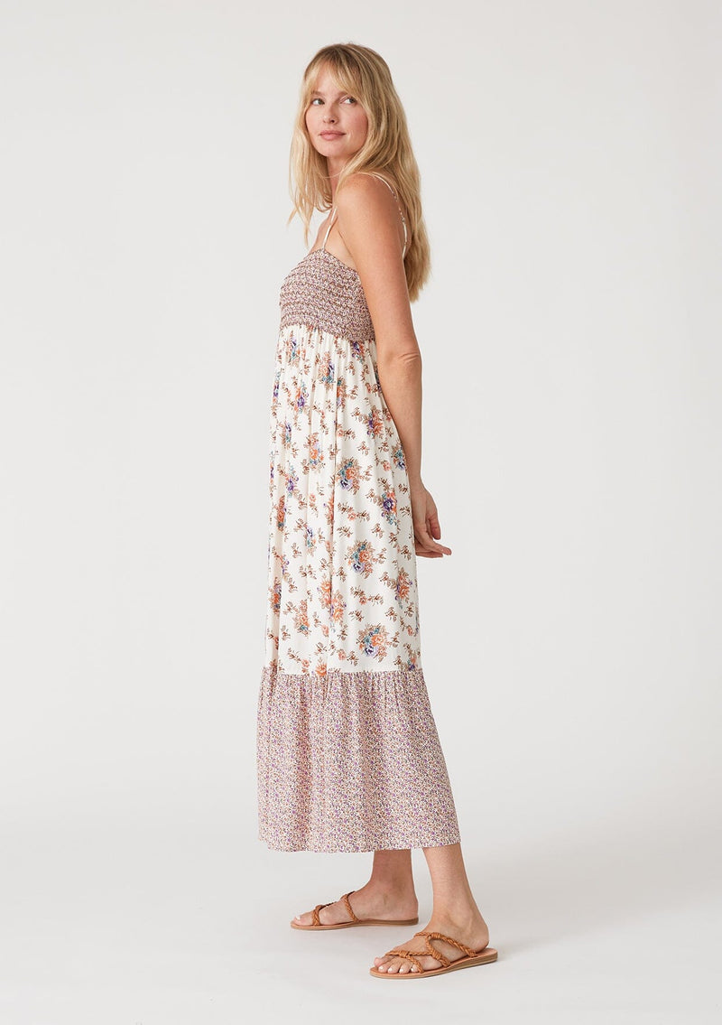 [Color: Natural/Purple] A side facing image of a blonde model wearing a best selling bohemian maxi dress in an off white and purple floral border print. With adjustable spaghetti straps, a square neckline, a smocked fitted bodice, a flowy tiered skirt, side pockets, and a button front. 