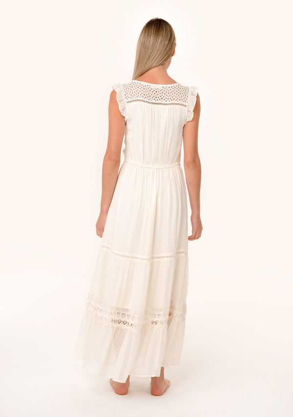 [Color: Vanilla] A back facing image of a blonde model wearing a flowy bohemian white maxi dress with short flutter sleeves, eyelet lace trim, embroidery, and a tassel tie drawstring waist.