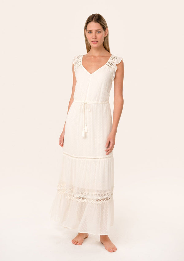 [Color: Vanilla] A front facing image of a blonde model wearing a flowy bohemian white maxi dress with short flutter sleeves, eyelet lace trim, embroidery, and a tassel tie drawstring waist.
