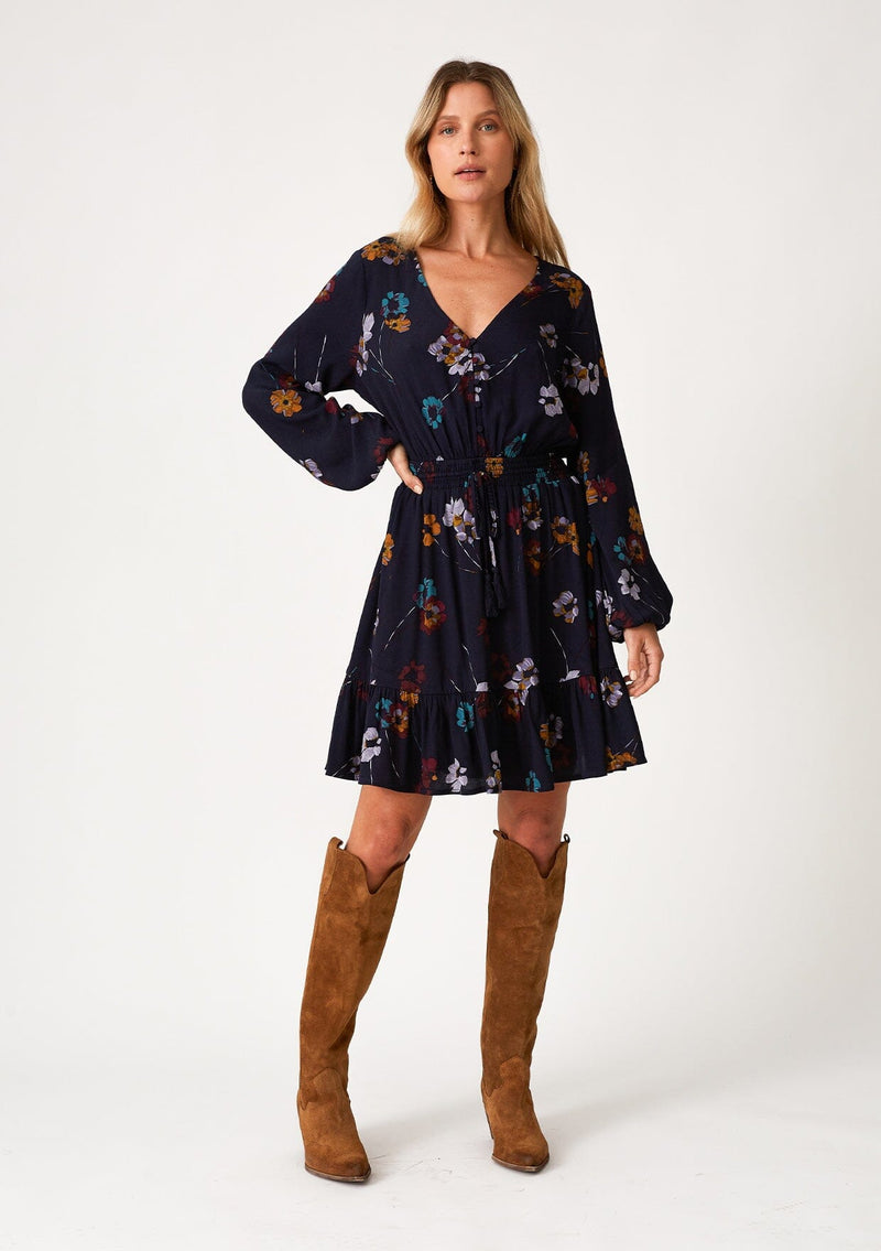 [Color: Navy/Mustard] A full body front facing image of a blonde model wearing a bohemian fall mini dress in a blue floral print. With long sleeves, a v neckline, a smocked elastic waist, a drawstring waist with tassel ties, a tiered mini skirt, and a self covered button front top. 