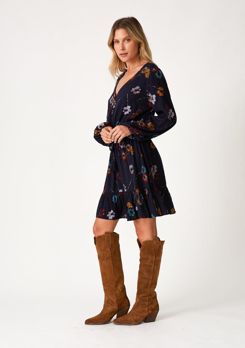 [Color: Navy/Mustard] A side facing image of a blonde model wearing a bohemian fall mini dress in a blue floral print. With long sleeves, a v neckline, a smocked elastic waist, a drawstring waist with tassel ties, a tiered mini skirt, and a self covered button front top. 