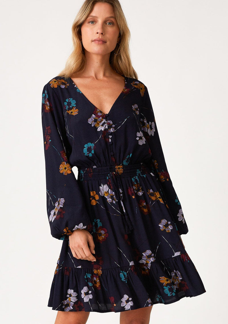 [Color: Navy/Mustard] A front facing image of a blonde model wearing a bohemian fall mini dress in a blue floral print. With long sleeves, a v neckline, a smocked elastic waist, a drawstring waist with tassel ties, a tiered mini skirt, and a self covered button front top. 