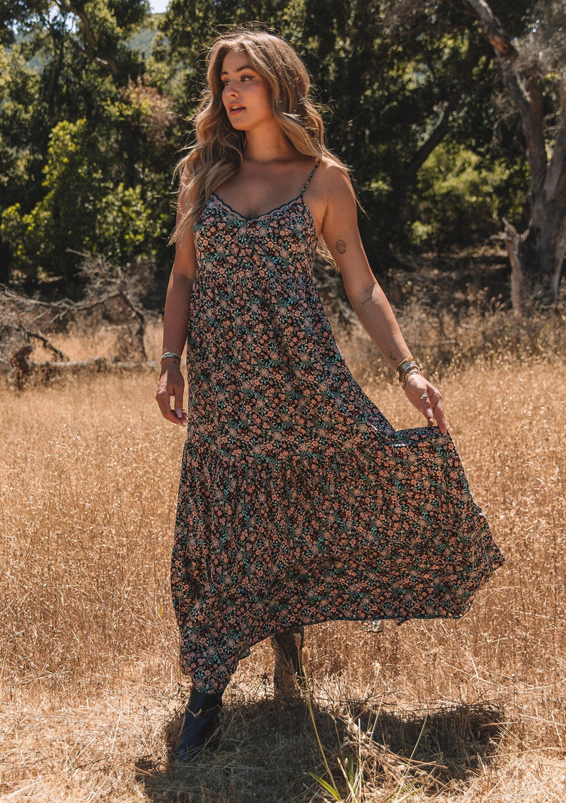 [Color: Black/Teal] A front facing image of a blonde model standing outside wearing a bohemian maxi dress in a black and teal floral print. With adjustable spaghetti straps, a scalloped trim v neckline with contrast thread details, a tiered flowy silhouette, side pockets, and an empire waist. 