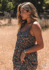 [Color: Black/Teal] A close up side facing image of a blonde model standing outside wearing a bohemian maxi dress in a black and teal floral print. With adjustable spaghetti straps, a scalloped trim v neckline with contrast thread details, a tiered flowy silhouette, side pockets, and an empire waist.