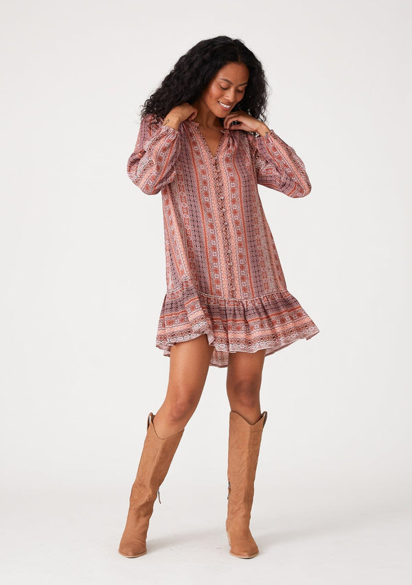 [Color: Cinnamon/Coral] A full body front facing image of a brunette model wearing a bohemian fall mini dress in a pink print. With long sleeves, a flowy baby doll silhouette, a tiered skirt, a self covered button front, and a v neckline. 