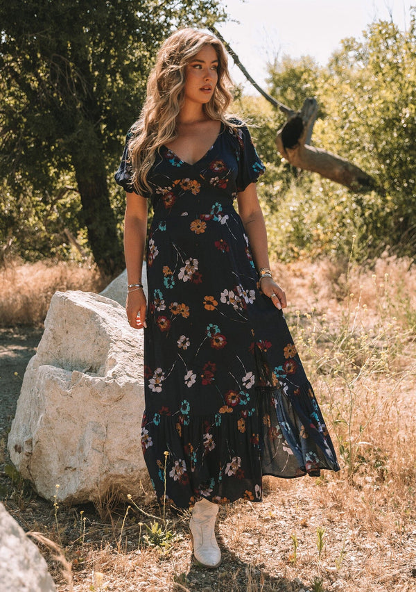 [Color: Navy/Mustard] A front facing image of a blonde model standing outside wearing a bohemian fall maxi dress in a navy blue and mustard yellow floral print. With short puff sleeves, a v neckline, a long flowy tiered skirt, and a side slit. 